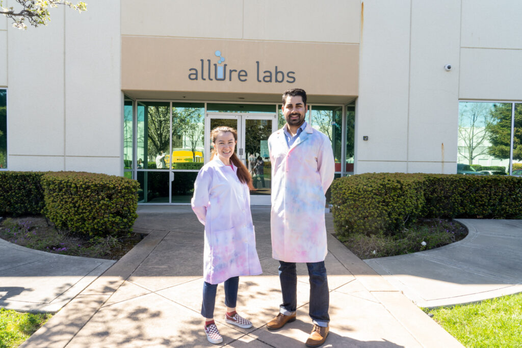 Two professionals wearing pastel tie-dye lab coats stand in front of the Allure Labs headquarters, a leading skincare contract manufacturer specializing in innovative formulations and beauty product development.