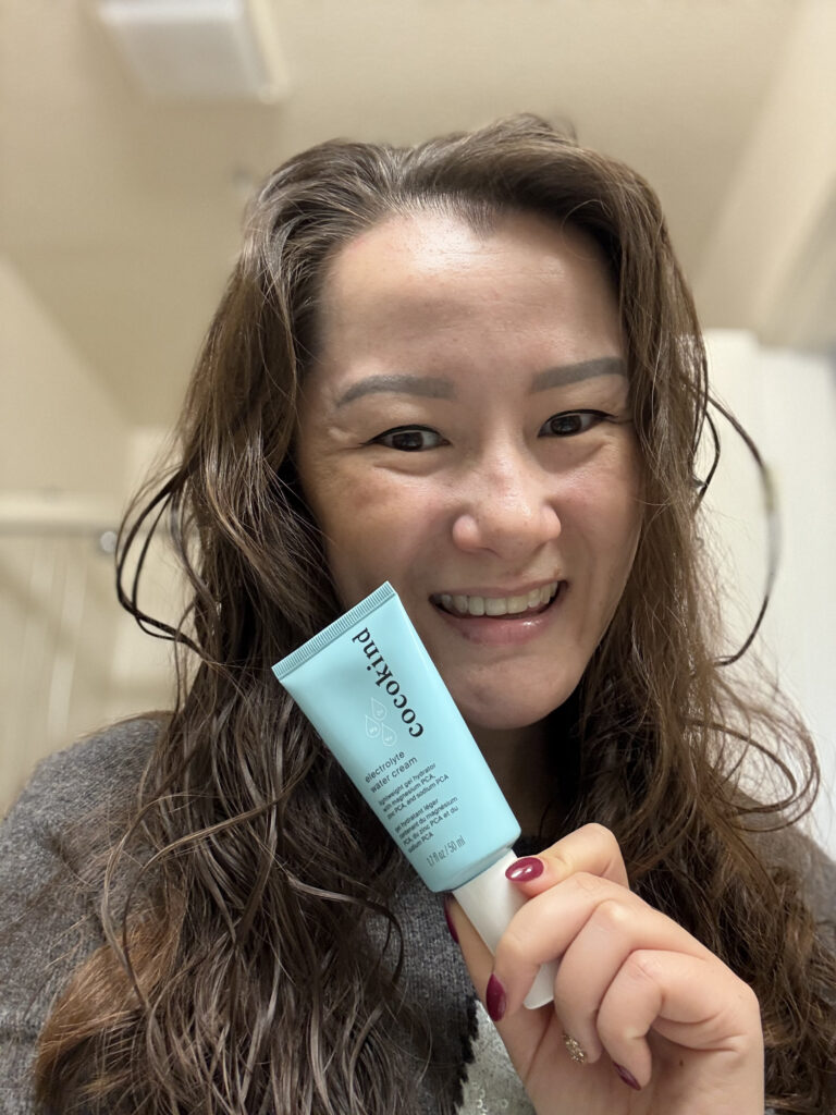 A smiling woman holding a tube of Cocokind Electrolyte Water Cream Moisturizer, showcasing Skin Positivity with Cocokind and highlighting its role in her skincare routine.