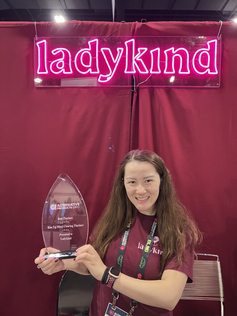 A smiling woman holds an award for 'Best Tincture' at the Alternative Products Expo, presented to Ladykind for their Rise Up Daily Mind Clearing Tincture, with a neon Ladykind sign in the background.